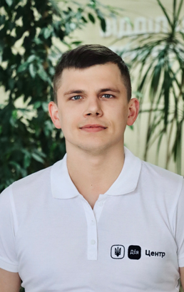 Чоловік у білій сорочці поло з логотипом стоїть на фоні зелених рослин в офісі.